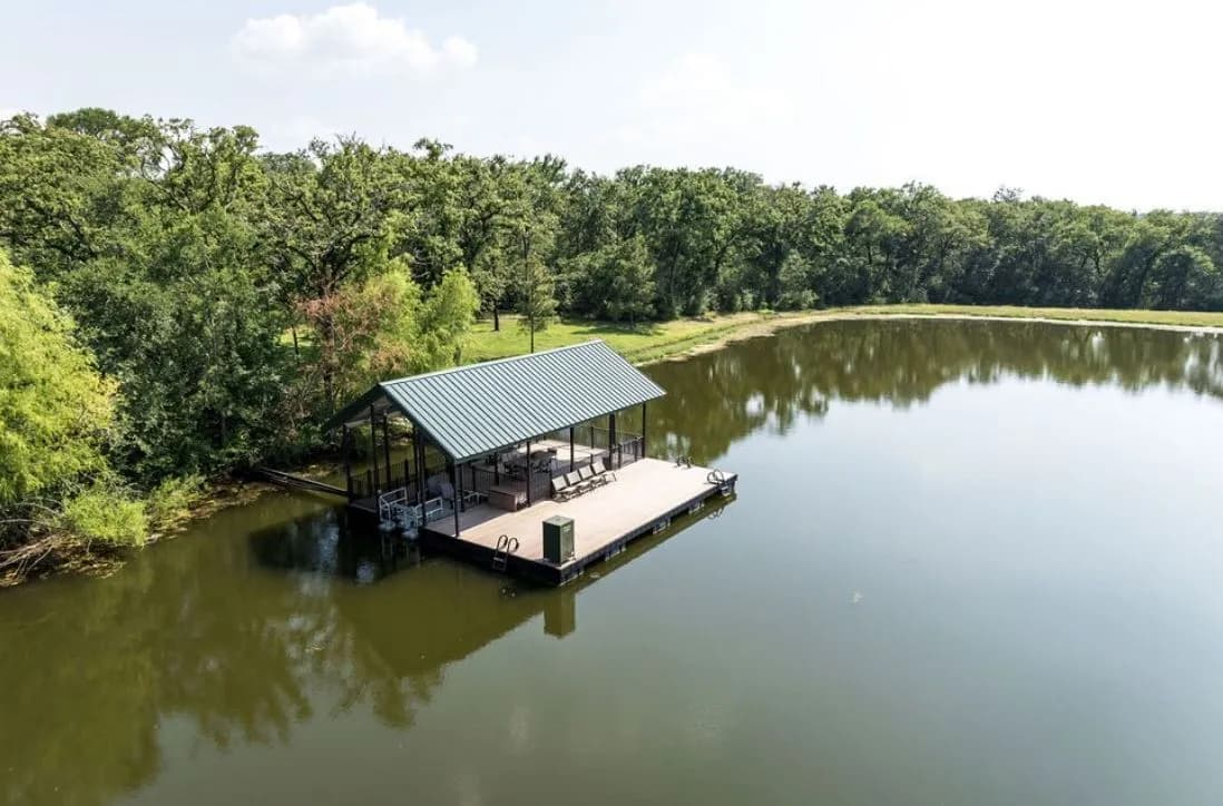 Aerial view of a covered aluminum floating dock with a green metal roof and composite deck, mirrored on a calm, tree-lined pond